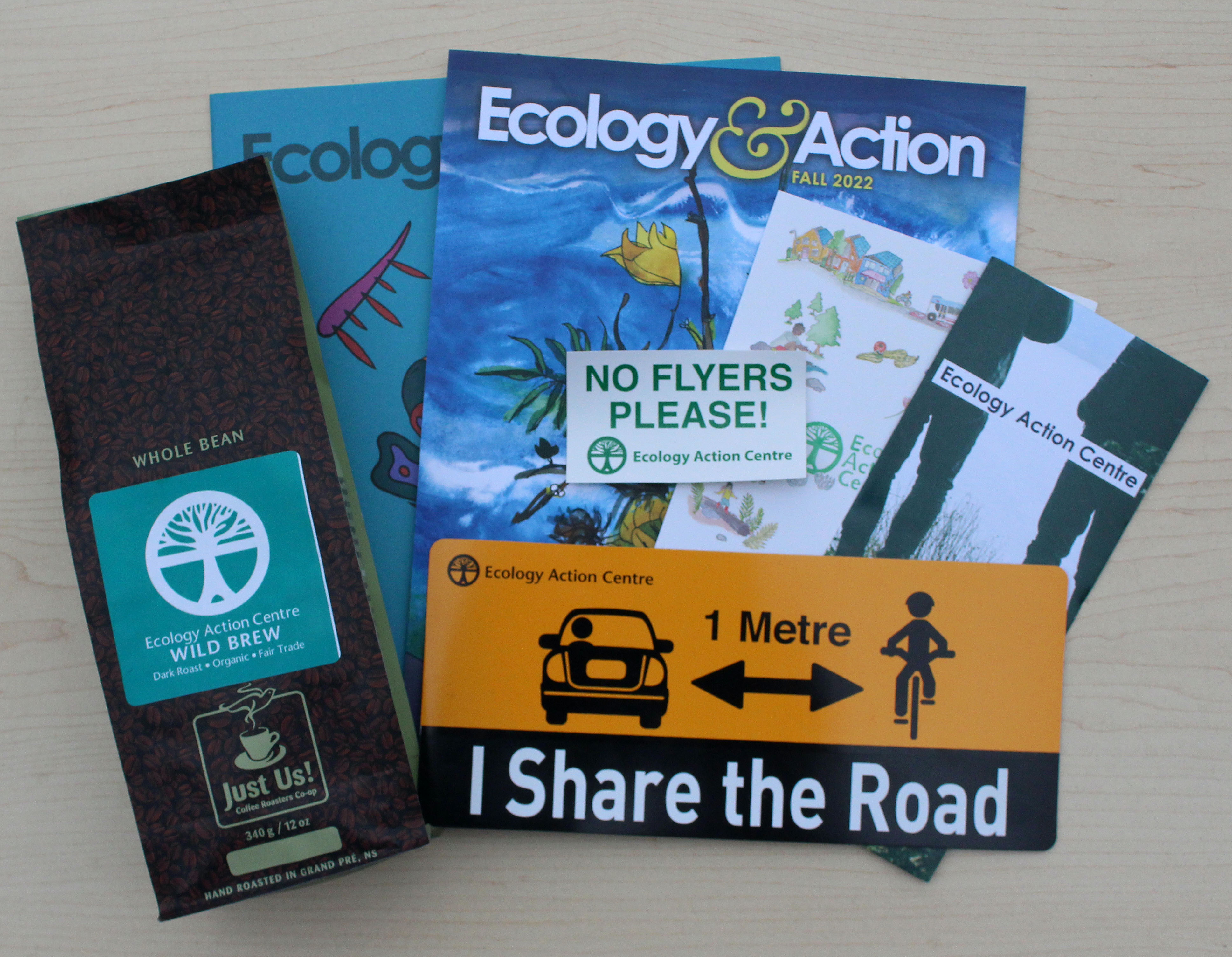 a bag of coffee, a brochure, two ecology action magazines, a no flyers sticker, a I share the road car magnet and a card laid out on a desk.