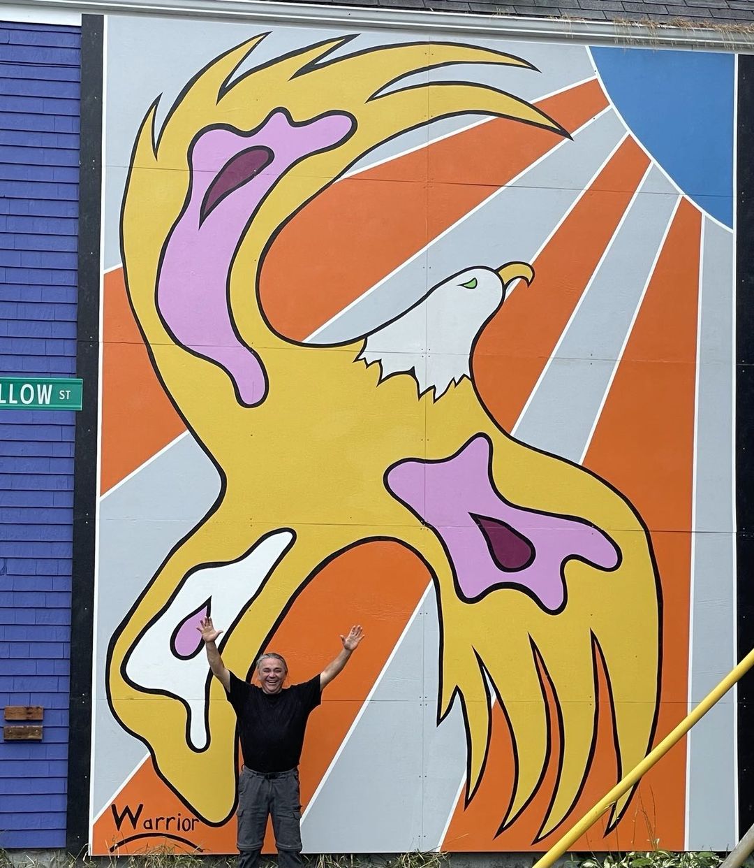 Lorne Julien stands in front of a brightly coloured mural depicting an eagle and a shining sun