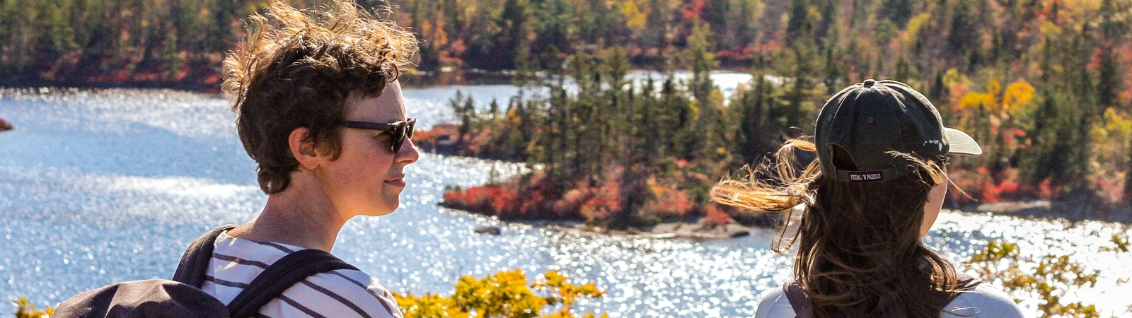 two people wearing backpacks stand at a point overlooking a lake and forest in autumn