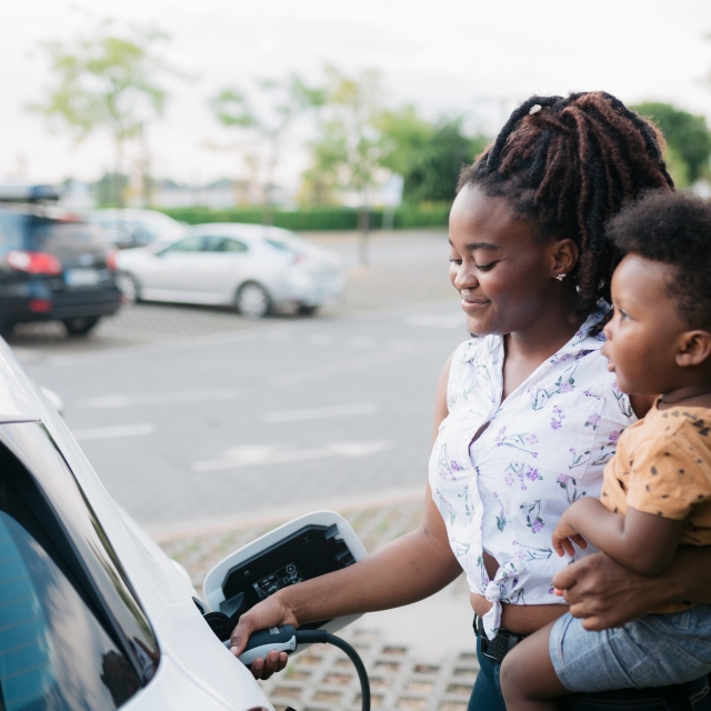 Young adult mother charging her electric car parked next to the charging station and holding her baby son in her other hand