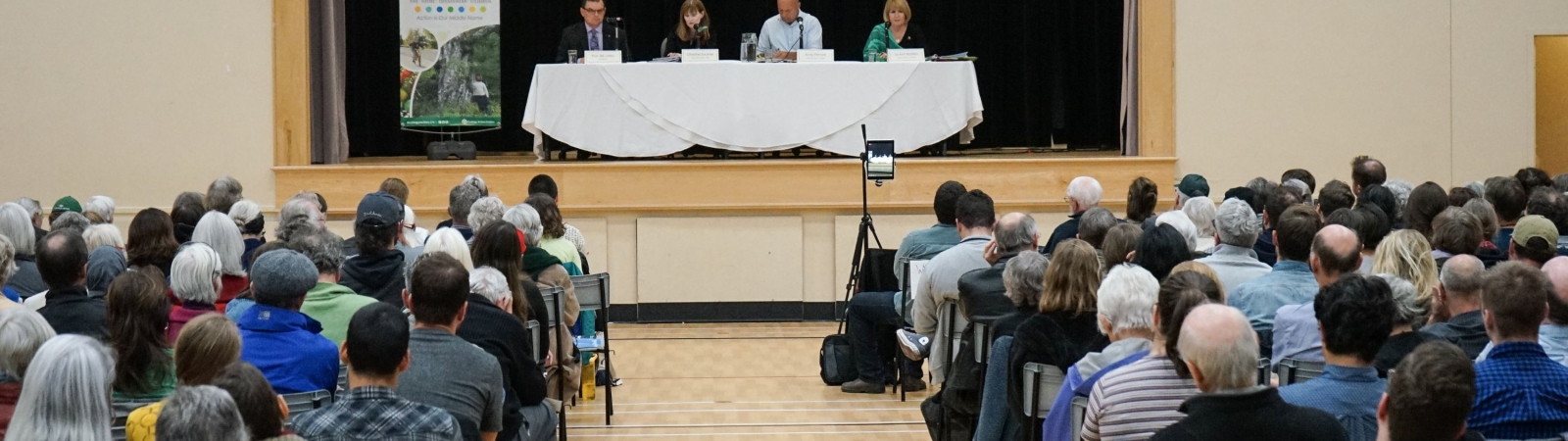 a large group of people watching an election debate with four people at a table  with a white table clothe on a stage.