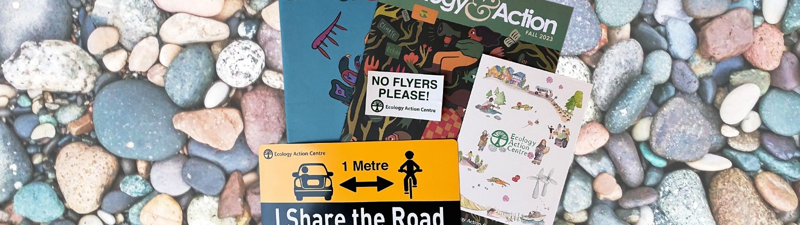 Contents of a gift membership on a backdrop of sea pebbles: two Ecology & Action magazines, a No Flyers Please sticker, an I Share the Road car magnet and an EAC card