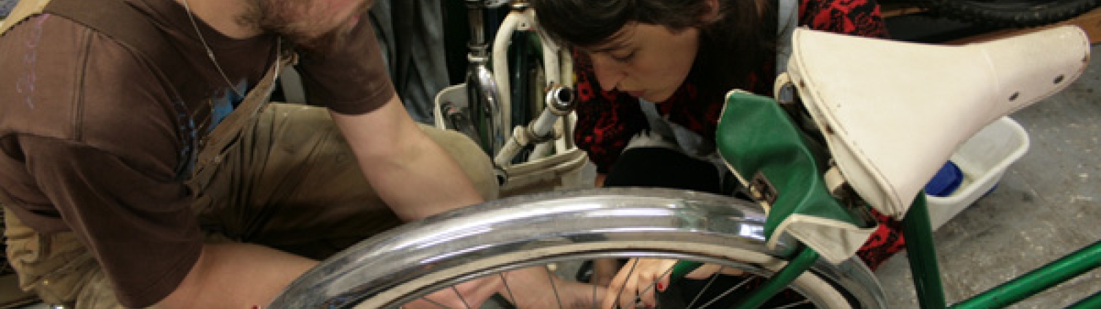 Two people are kneeling and wrenching the back wheel of a bicycle together in the Bike Again workshop