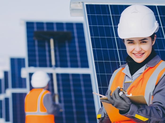 A women holding a clip board wearing a hard hat standing in front of solar panels.