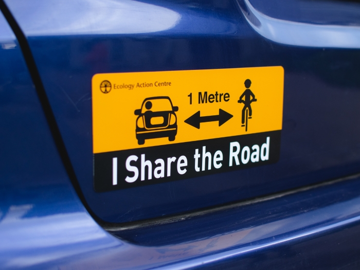 A yellow car magnet on a blue bumper of a car that reads I Share The Road in black letters and a drawing of a cyclist in black