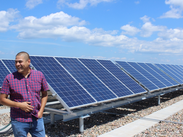 a smiling bald man wearing a pink and purple checkered shirt stands in front of rows of solar panels on a gravel surface