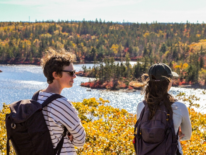 two people wearing backpacks stand at a point overlooking a lake and forest in autumn